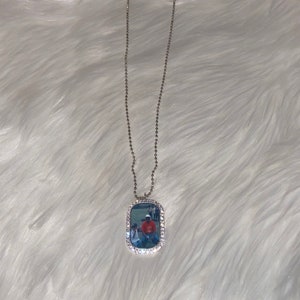 Könnte beinhalten: Silberfarbene Halskette mit rechteckigem Anhänger. Der Anhänger zeigt ein Foto, umgeben von einem Rand aus kleinen, klaren Kristallen. Die Halskette hat eine feine Kugelkette und hängt auf einer weißen, flauschigen Oberfläche.