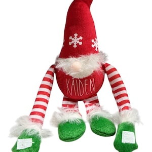 May include: A Christmas gnome doll with a red pointed hat decorated with white snowflakes. The gnome has a white beard, a red body with the name "KAIDEN", and red and white striped arms and legs. The feet are green with white trim.
