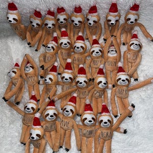 May include: A collection of plush sloths wearing red Santa hats. Each sloth has a name tag. The sloths are light brown with white faces and black eyes. Some sloths have their arms raised. The background is a white, fluffy surface.