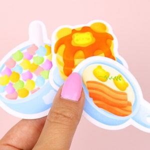 Puede incluir: Tres pegatinas de comida para el desayuno de dibujos animados: un taz&oacute;n de cereal de colores, una pila de panqueques con jarabe naranja y una sart&eacute;n de tocino con dos huevos amarillos.