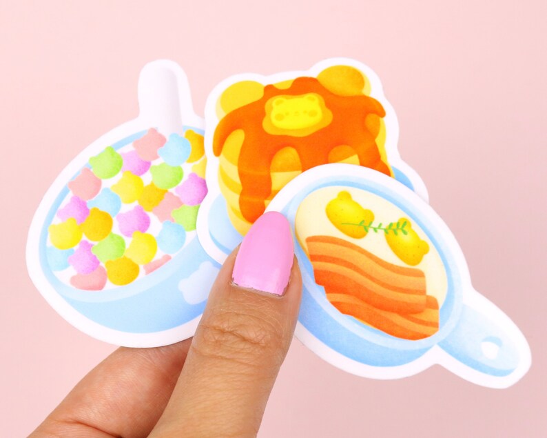 Puede incluir: Tres pegatinas de comida para el desayuno de dibujos animados: un taz&oacute;n de cereal de colores, una pila de panqueques con jarabe naranja y una sart&eacute;n de tocino con dos huevos amarillos.