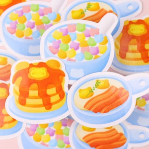 Puede incluir: Un conjunto de pegatinas coloridas que representan diferentes alimentos para el desayuno, incluyendo panqueques, cereales y huevos con tocino. Las pegatinas est&aacute;n ilustradas en un estilo de dibujos animados con un fondo azul claro.