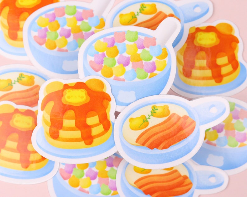 Puede incluir: Un conjunto de pegatinas coloridas que representan diferentes alimentos para el desayuno, incluyendo panqueques, cereales y huevos con tocino. Las pegatinas est&aacute;n ilustradas en un estilo de dibujos animados con un fondo azul claro.