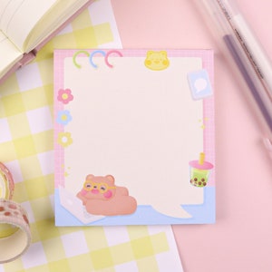 May include: A pink and blue notepad featuring a cartoon bear wearing glasses and using a laptop. The notepad has a speech bubble with a cartoon boba tea cup and a cartoon bear with a crown. The notepad is on a yellow and white checkered background.