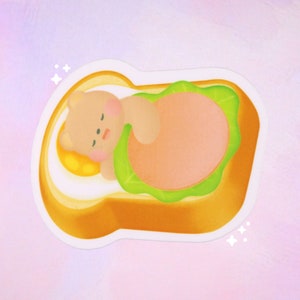 May include: A cartoon bear sleeping inside a slice of bread with a fried egg and lettuce. The bread is yellow and the lettuce is green.