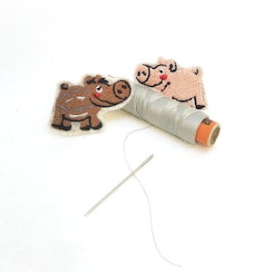 May include: Two embroidered animal patches, a brown boar and a pink pig, sit near a spool of gray thread and a needle. The boar has brown and gray details, while the pig has red cheeks. The needle is silver.