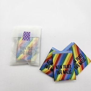 Peut inclure: Un ensemble d'étiquettes en tissu rectangulaires avec un motif de rayures arc-en-ciel en diagonale et le texte noir "KEIN PLATZ FÜR NAZIS". Trois étiquettes sont libres et une est dans une enveloppe en papier blanc translucide scellée.