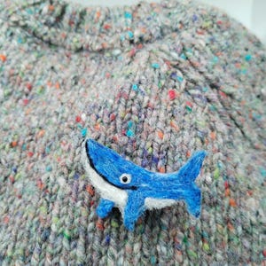 May include: A needle-felted blue whale brooch with a white underbelly and a single black eye, attached to a grey knit sweater featuring a multi-colored flecked pattern throughout the fabric, creating a textured appearance on the garment.