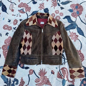 Puede incluir: Chaqueta de ante marrón con cremallera, con un diseño de patchwork de rombos en tonos marrón y burdeos en el cuello, el pecho y los puños. La chaqueta tiene dos bolsillos delanteros y está sobre una tela estampada floral.