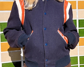 May include: Navy blue varsity jacket with orange and white accents on the shoulders and pockets. The jacket has a ribbed collar, cuffs, and hem. It features blue buttons down the front and is a classic outerwear piece.