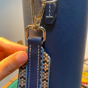 May include: A close-up of a blue bag with a gold metal clasp and a patterned strap. The strap is made of woven fabric with brown, white, and blue colors.