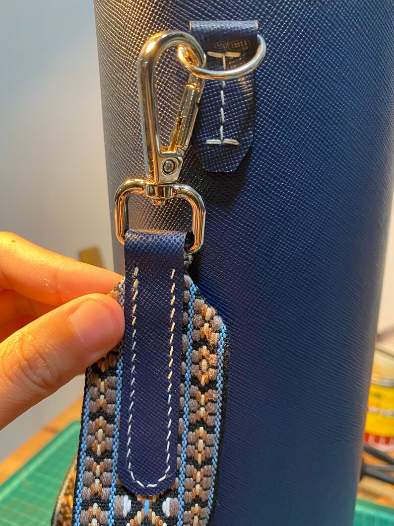 May include: A close-up of a blue bag with a gold metal clasp and a patterned strap. The strap is made of woven fabric with brown, white, and blue colors.
