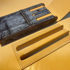 K&ouml;nnte beinhalten: Ein handgefertigtes Leder-Stiftetui mit einem verwitterten grau-schwarzen Finish, wei&szlig;en N&auml;hten und einem beigefarbenen Innenraum. Das Etui ist ge&ouml;ffnet und zeigt einen beigefarbenen Ledereinsatz mit zwei erh&ouml;hten Trennw&auml;nden. Das Etui liegt auf einer beigefarbenen Oberfl&auml;che.
