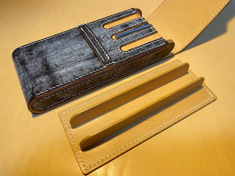K&ouml;nnte beinhalten: Ein handgefertigtes Leder-Stiftetui mit einem verwitterten grau-schwarzen Finish, wei&szlig;en N&auml;hten und einem beigefarbenen Innenraum. Das Etui ist ge&ouml;ffnet und zeigt einen beigefarbenen Ledereinsatz mit zwei erh&ouml;hten Trennw&auml;nden. Das Etui liegt auf einer beigefarbenen Oberfl&auml;che.
