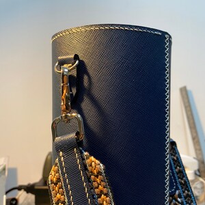 May include: A dark blue leather bag with a gold clasp and a patterned strap. The bag has white stitching around the edges.