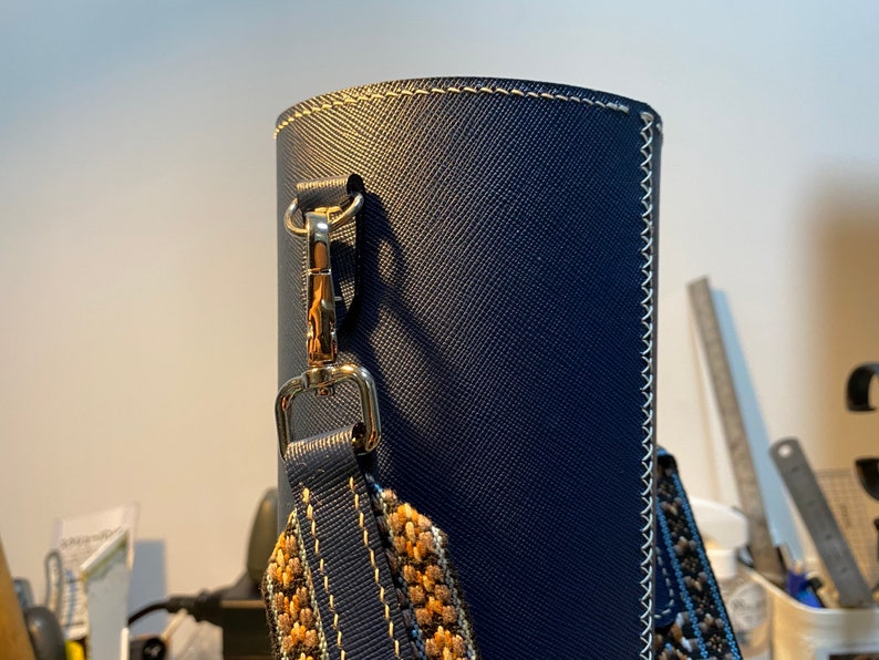 May include: A dark blue leather bag with a gold clasp and a patterned strap. The bag has white stitching around the edges.