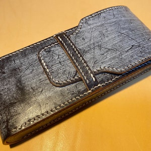 K&ouml;nnte beinhalten: Eine handgefertigte Ledertasche in Grau- und Schwarzt&ouml;nen mit strukturierter Oberfl&auml;che und wei&szlig;en N&auml;hten. Die Tasche hat einen Klappverschluss mit passendem Lederriemen und ist f&uuml;r einen kleinen Gegenstand konzipiert.
