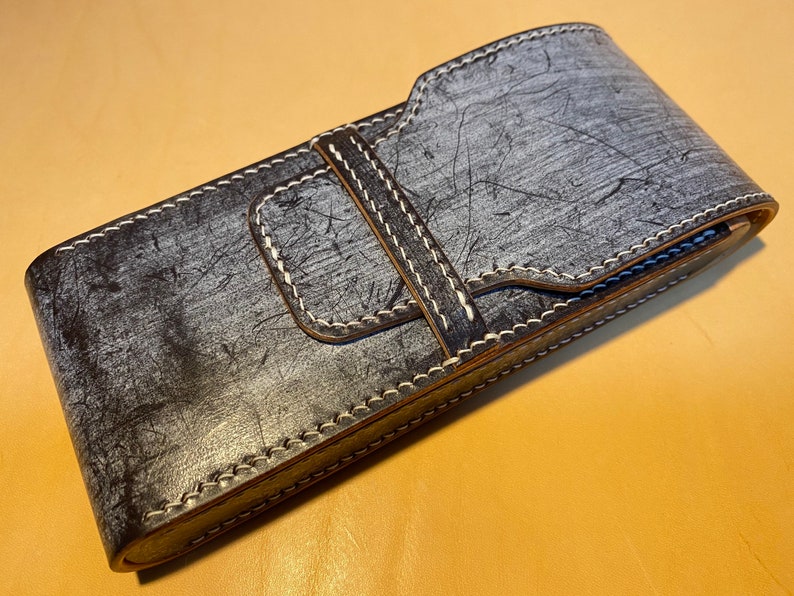 K&ouml;nnte beinhalten: Eine handgefertigte Ledertasche in Grau- und Schwarzt&ouml;nen mit strukturierter Oberfl&auml;che und wei&szlig;en N&auml;hten. Die Tasche hat einen Klappverschluss mit passendem Lederriemen und ist f&uuml;r einen kleinen Gegenstand konzipiert.