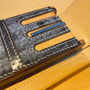 K&ouml;nnte beinhalten: Nahaufnahme eines handgefertigten Leder-Stiftetuis. Das Etui hat eine verwitterte grau-schwarze Au&szlig;enseite mit wei&szlig;en N&auml;hten und hellbraunen Lederakzenten. Ein blauer Metallakzent ist sichtbar. Das Etui ist ge&ouml;ffnet und zeigt ein hellbraunes Innenfutter.