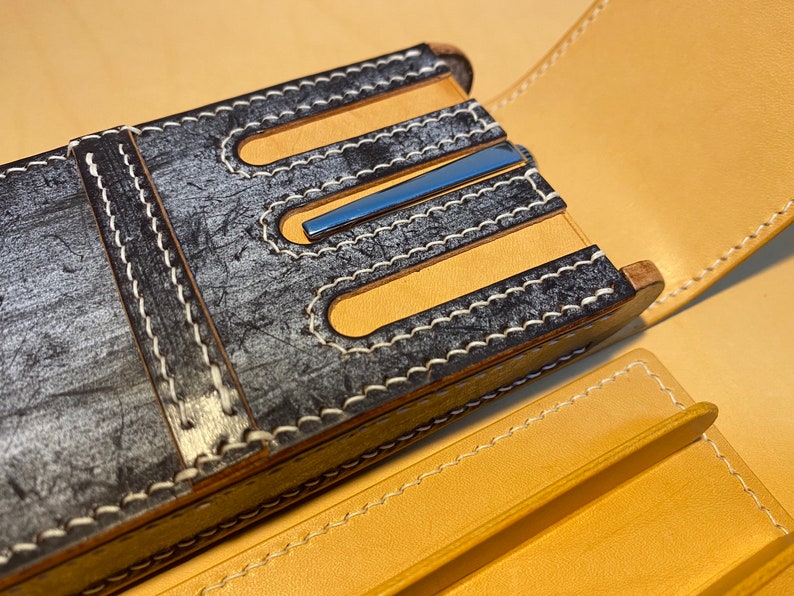 K&ouml;nnte beinhalten: Nahaufnahme eines handgefertigten Leder-Stiftetuis. Das Etui hat eine verwitterte grau-schwarze Au&szlig;enseite mit wei&szlig;en N&auml;hten und hellbraunen Lederakzenten. Ein blauer Metallakzent ist sichtbar. Das Etui ist ge&ouml;ffnet und zeigt ein hellbraunes Innenfutter.