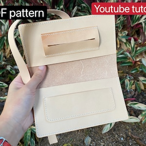 Può includere: Un portafoglio in pelle marrone chiaro con una patta e un cinturino. Il portafoglio ha una tasca per le banconote e uno slot per le carte. Il portafoglio è aperto e l'interno è visibile. Il portafoglio è su uno sfondo verde.
