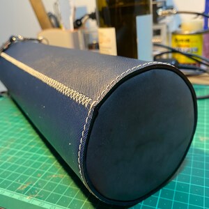 May include: A dark blue cylindrical carrying case with a white stitched seam. The case is made of a textured material and has a smooth, rounded end.