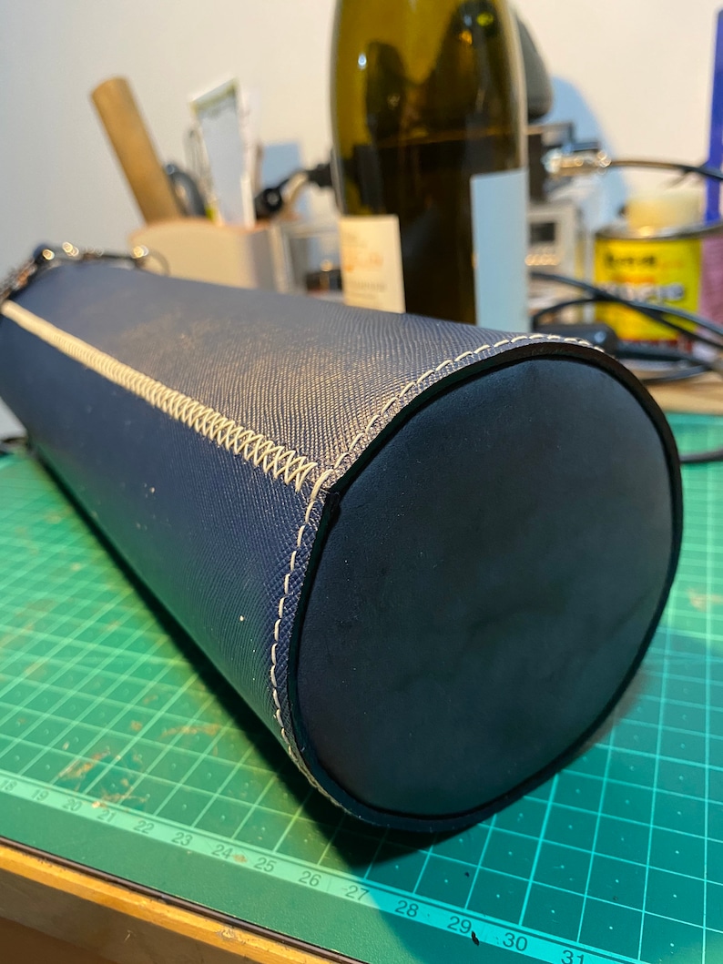 May include: A dark blue cylindrical carrying case with a white stitched seam. The case is made of a textured material and has a smooth, rounded end.