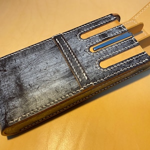 K&ouml;nnte beinhalten: Rechteckige Darttasche aus Leder mit einem Used-Look in Grau und Schwarz. Die Tasche hat wei&szlig;e N&auml;hte und beige Lederakzente. Das Innere hat einen blauen Metallakzent. Ca. 18 cm lang.