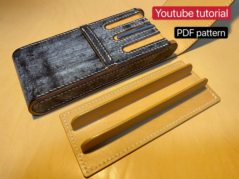 K&ouml;nnte beinhalten: Ein handgefertigtes Leder-Stifteetui in Grau mit hellbraunen Akzenten und wei&szlig;en N&auml;hten. Das Etui hat einen Klappverschluss. Ein separates hellbraunes Lederst&uuml;ck mit zwei erh&ouml;hten Trennw&auml;nden ist ebenfalls abgebildet. Text: Youtube-Tutorial, PDF-Muster.