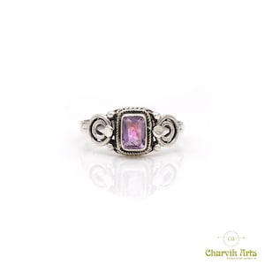 May include: A silver ring with a rectangular, light purple gemstone at the center. The ring has a decorative design with swirls on either side of the gemstone. The ring is set against a white background.