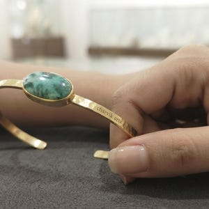 Könnte beinhalten: Ein goldfarbenes Metallarmband mit einem glatten, ovalen, grün-schwarz marmorierten Stein in einer Fassung. Das Band hat eine gehämmerte Textur und der Schriftzug charvik arts ist seitlich in der Nähe der Steinfassung eingraviert.