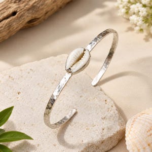 Op de afbeelding: Een zilverkleurige metalen manchetarmband met een gehamerde textuur langs de band en een enkele witte kaurischelp in het midden aan de voorzijde. De armband heeft een open ontwerp, waardoor deze gemakkelijk om de pols kan worden geschoven.