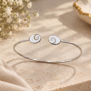 Op de afbeelding: Een zilverkleurige open armband met twee ovale uiteinden. Elk uiteinde is ingelegd met een witte Shiva-oogschelp met een natuurlijk grijs spiraalpatroon. De band heeft een getextureerde, metalen afwerking en een slank, flexibel ontwerp.