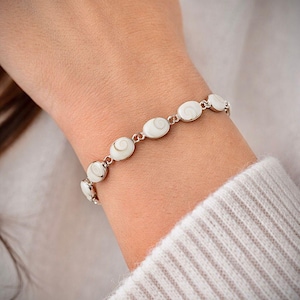 Puede incluir: Un brazalete plateado con segmentos ovalados de concha blanca con diseño en espiral. El brazalete está conectado por pequeños eslabones plateados y se lleva en la muñeca, una delicada pieza de joyería.