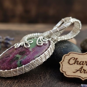 Puede incluir: Un colgante envuelto en alambre con una piedra preciosa de rubí zoisita de forma ovalada con inclusiones de color magenta intenso y verde. La piedra está encerrada en un intrincado trabajo de alambre plateado que forma un asa decorativa.