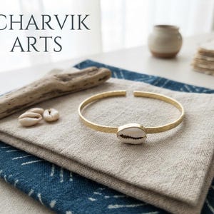 Może przedstawiać: Złota metalowa bransoletka typu mankiet z pojedynczą białą muszlą kauri pośrodku. Tekst CHARVIK ARTS jest widoczny w lewym górnym rogu. Bransoletka ma młotkowaną fakturę i otwarty design, zaprezentowana wraz z małymi muszelkami.