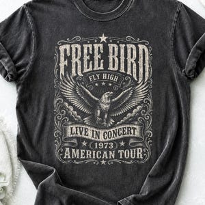 May include: Dark gray t-shirt featuring a distressed graphic with the words "FREE BIRD" above an eagle with outstretched wings. Below the eagle, the text reads "LIVE IN CONCERT 1973 AMERICAN TOUR". The shirt has a vintage, worn look.