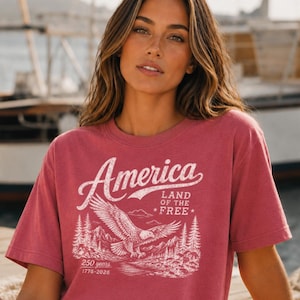 May include: A dusty rose short-sleeve t-shirt featuring a white graphic print of a bald eagle in flight over a mountain landscape. The text reads America, Land of the Free, and 250 years 1776-2026 in a distressed, vintage-style font design.