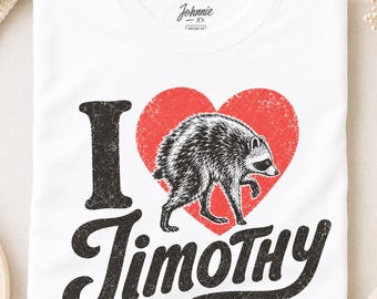 May include: A white short-sleeved t-shirt featuring a graphic print with the text I LOVE TIMOTHY in black, distressed lettering. A red heart replaces the word LOVE, containing a detailed black and white illustration of a raccoon.