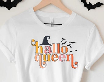 Op de afbeelding: Wit t-shirt met de tekst "Hallo Queen" in oranje, geel en roze, met een heksenhoed en vleermuizen. Het shirt hangt aan een houten hanger, met zwarte vleermuisuitsparingen erboven.