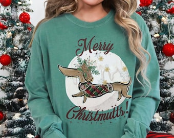 Peut inclure: Un sweat-shirt vert sarcelle avec les mots "Merry Christmutts!" et un teckel portant un pull à carreaux. Le chien est placé sur un cercle blanc avec des accents floraux. La personne porte un pantalon de pyjama à carreaux rouges et noirs.