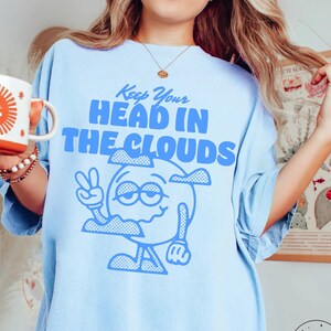 Puede incluir: Camiseta azul claro con un personaje de dibujos animados y el texto "Keep Your Head In The Clouds".