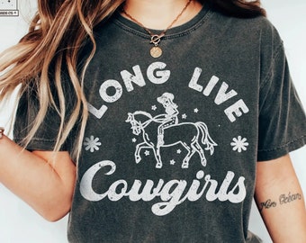 Puede incluir: Una camiseta gris oscura con un diseño gráfico blanco que dice "Long Live Cowgirls" con una vaquera montando un caballo. El gráfico está rodeado de estrellas.