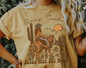 Könnte beinhalten: Senfgelbes T-Shirt mit einer Grafik einer Person, die in einer Wüstenlandschaft reitet. Die Texte "Sorry Cowboys" und "Jesus Has My Heart" sind aufgedruckt. Die Ärmel sind hochgekrempelt.