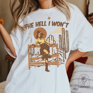 May include: White t-shirt with a graphic of a woman in a cowboy hat and boots, standing on a fence with a lasso. The text "THE HELL I WON'T" is printed above the graphic.