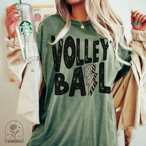 May include: A green t-shirt with the words "VOLLEYBALL" in black with a distressed look. A leopard print lightning bolt is in the center of the word "BALL".