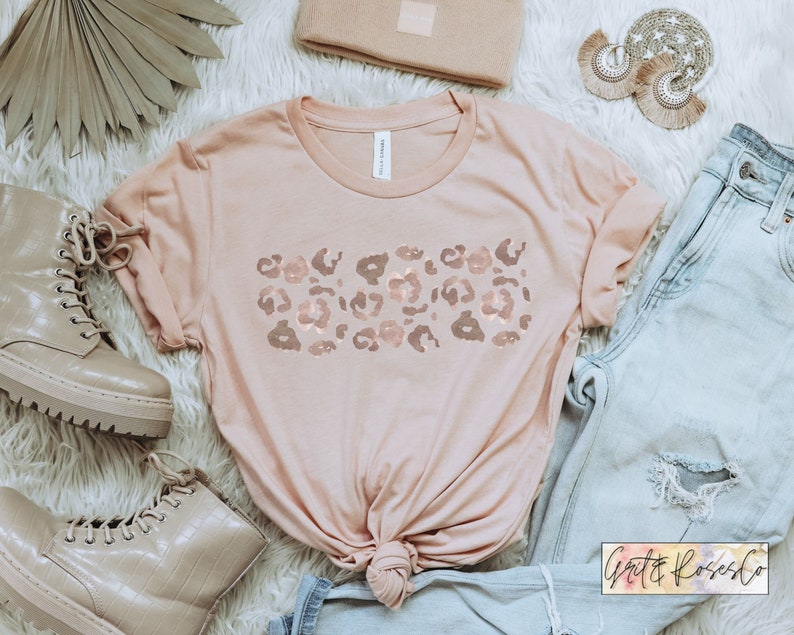 May include: A light pink t-shirt with a rose gold leopard print design. The shirt is styled with a knot in the front and is laid on a white fuzzy surface with a pair of light brown boots, a pair of light blue jeans, and a small brown wallet.