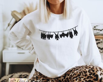 Könnte beinhalten: Weißer Pullover mit einer schwarzen Grafik von Fledermäusen, die an einer Wäscheleine hängen. Die Fledermäuse sind alle unterschiedlich groß und geformt.