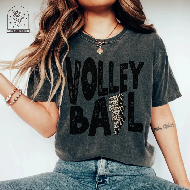 May include: A gray t-shirt with the text "VOLLEYBALL" in black with a leopard print lightning bolt graphic. The shirt is worn by a person with long brown hair.