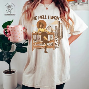 May include: A beige t-shirt with the text "The Hell I Won't" printed on it. The graphic features a woman in a cowboy hat and boots, standing on a fence with a lasso in her hand. The background is a desert scene with cacti.